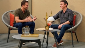 Two people seated on wicker chairs, a table with a lamp and a pitcher, and a backdrop of trees with purple flowers.