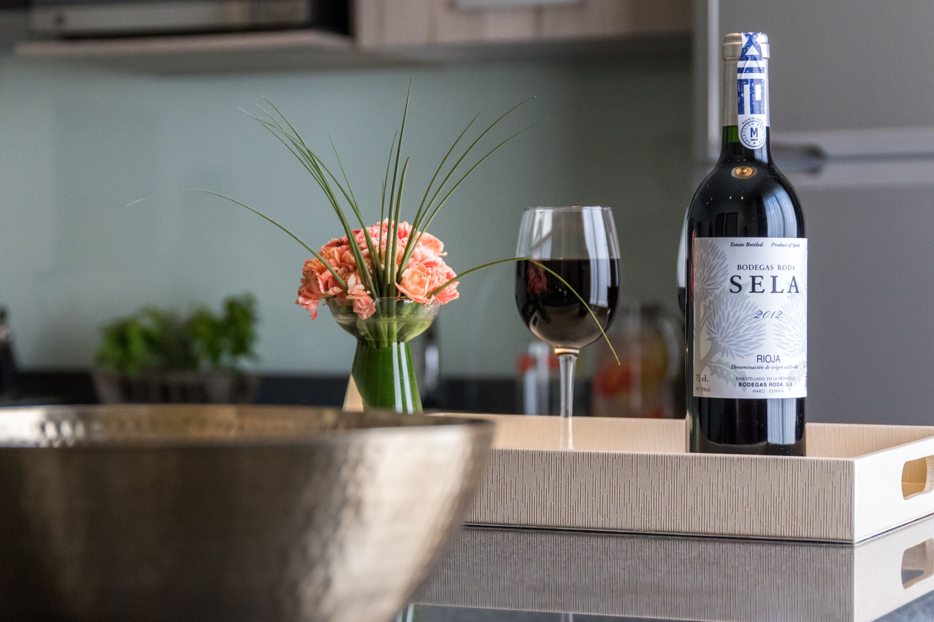 Una botella de vino Bodega Roda Sela, una copa de vino tinto y un florero con flores sobre una superficie de madera.
