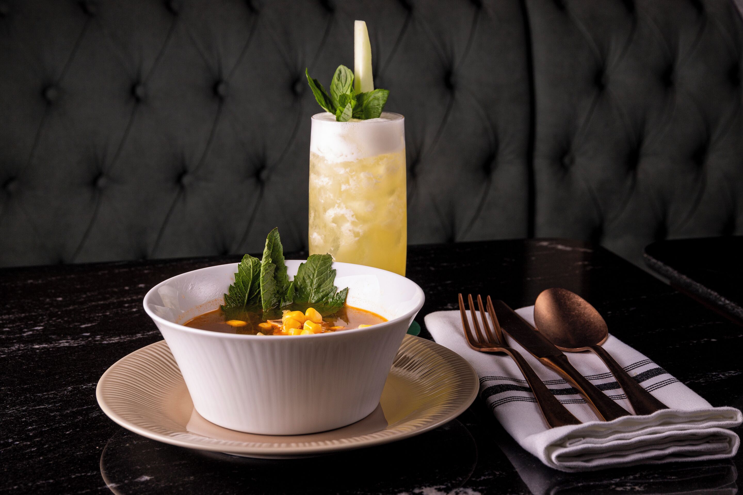 A bowl of soup with corn and mint garnish on a plate, accompanied by a tall glass of iced tea with a mint garnish, set on a black table with a tufted backrest.