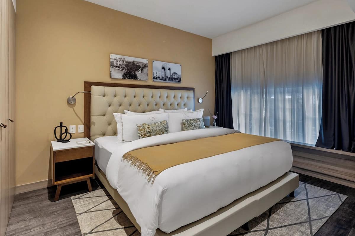 A hotel room with a large bed, a nightstand, a wall-mounted lamp, and two framed pictures above the bed.