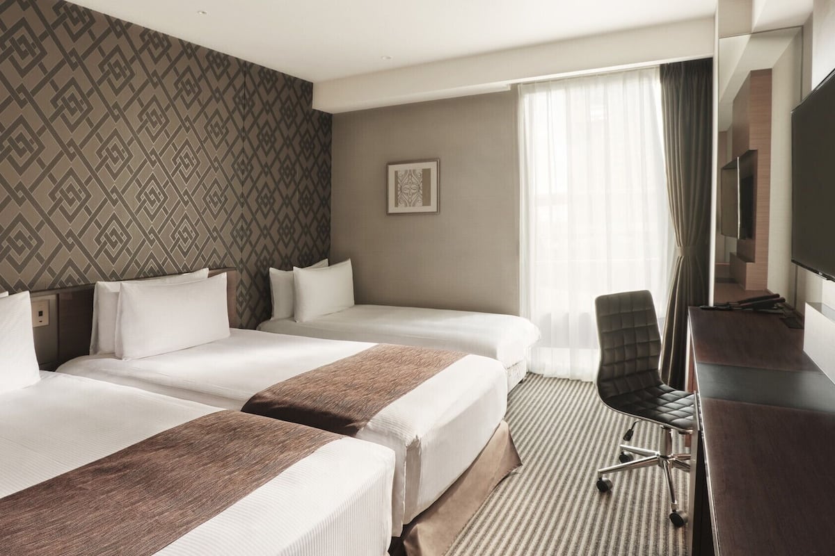 Hotellihuone, jossa on kaksi sänkyä, työpöytä, tuoli, televisio ja kuviollinen tapetti.