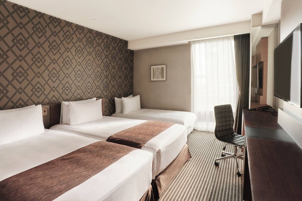 Hotellihuone, jossa on kaksi sänkyä, työpöytä, tuoli, televisio ja kuviollinen seinä.