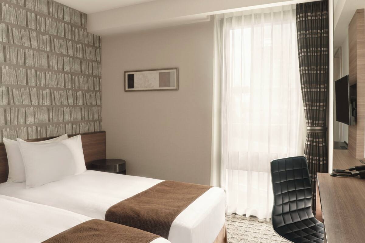Hotellihuone, jossa on suuri sänky, työpöytä tuolilla, televisio ja ikkuna, jossa on ohuet verhot.