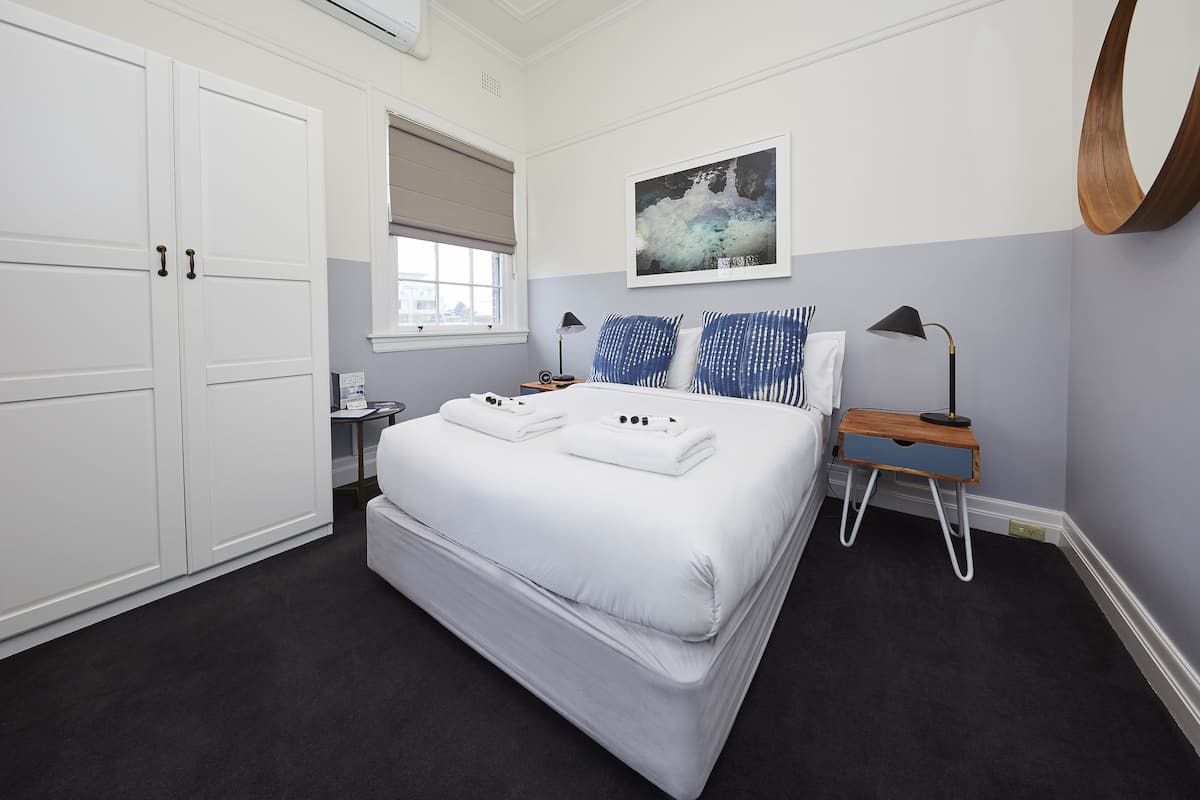 A bedroom with a large bed, a bedside table, a nightstand, a window with a view, and a painting on the wall.