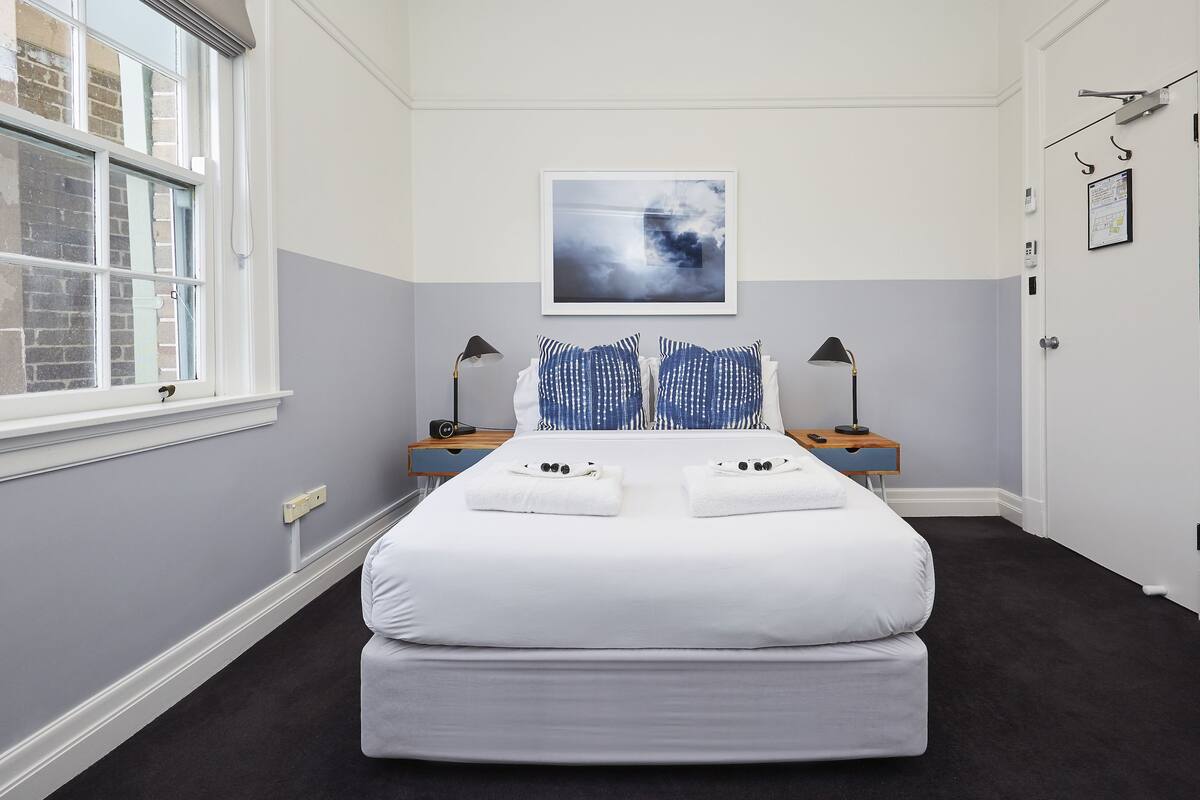 A neatly arranged bedroom with a large bed, a bedside table with a lamp, a framed picture on the wall, and a window with a view of the outside.