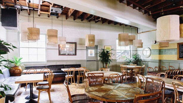 A spacious dining area with multiple wooden tables and chairs, a large potted plant, and a modern ceiling with exposed beams. - Bridgeview Hotel Willoughby (Willoughby)