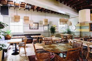 A spacious dining area with multiple wooden tables and chairs, a large potted plant, and a modern ceiling with exposed beams. - Bridgeview Hotel Willoughby (Willoughby)
