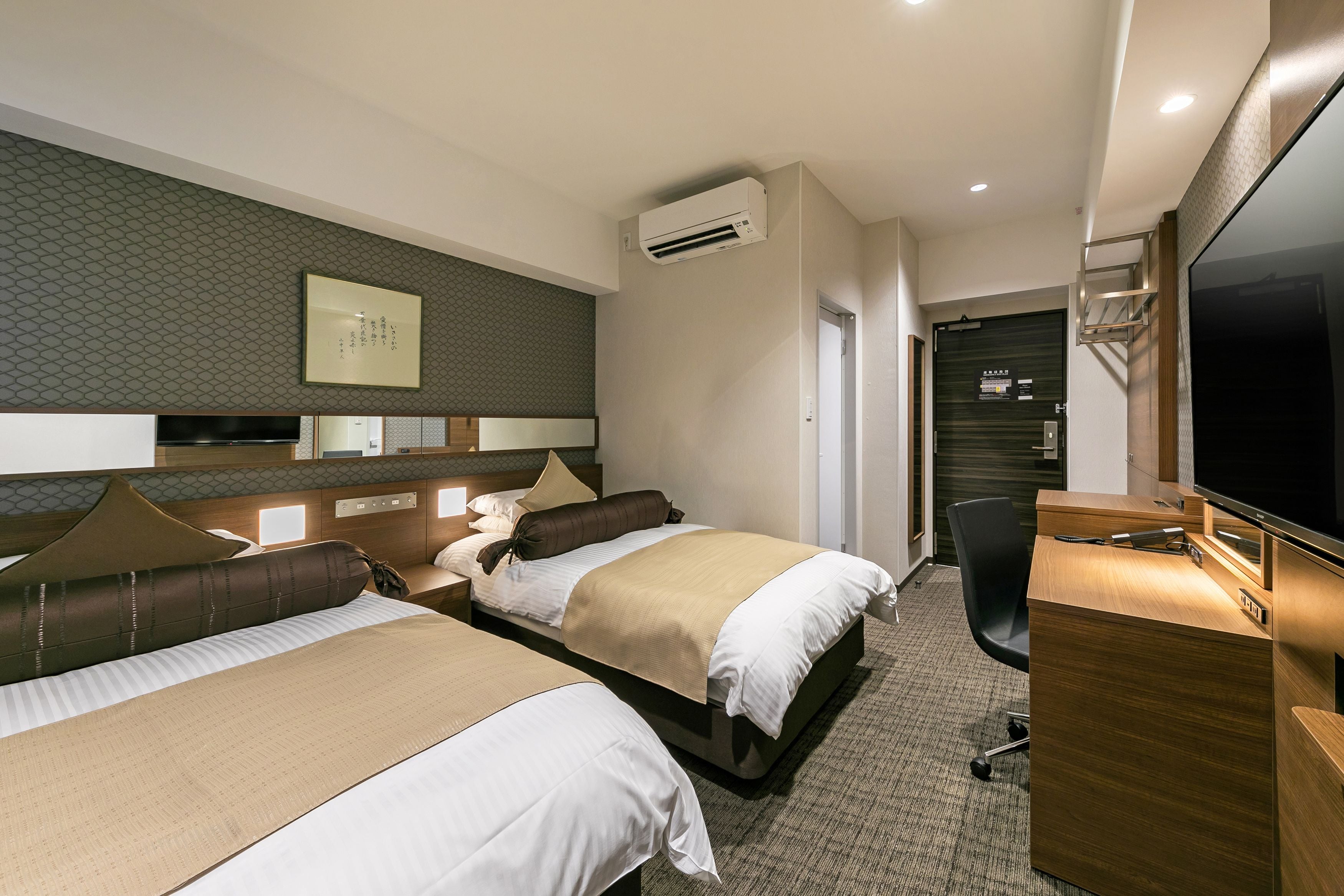 A hotel room with two beds, a desk, a TV, and an air conditioning unit.