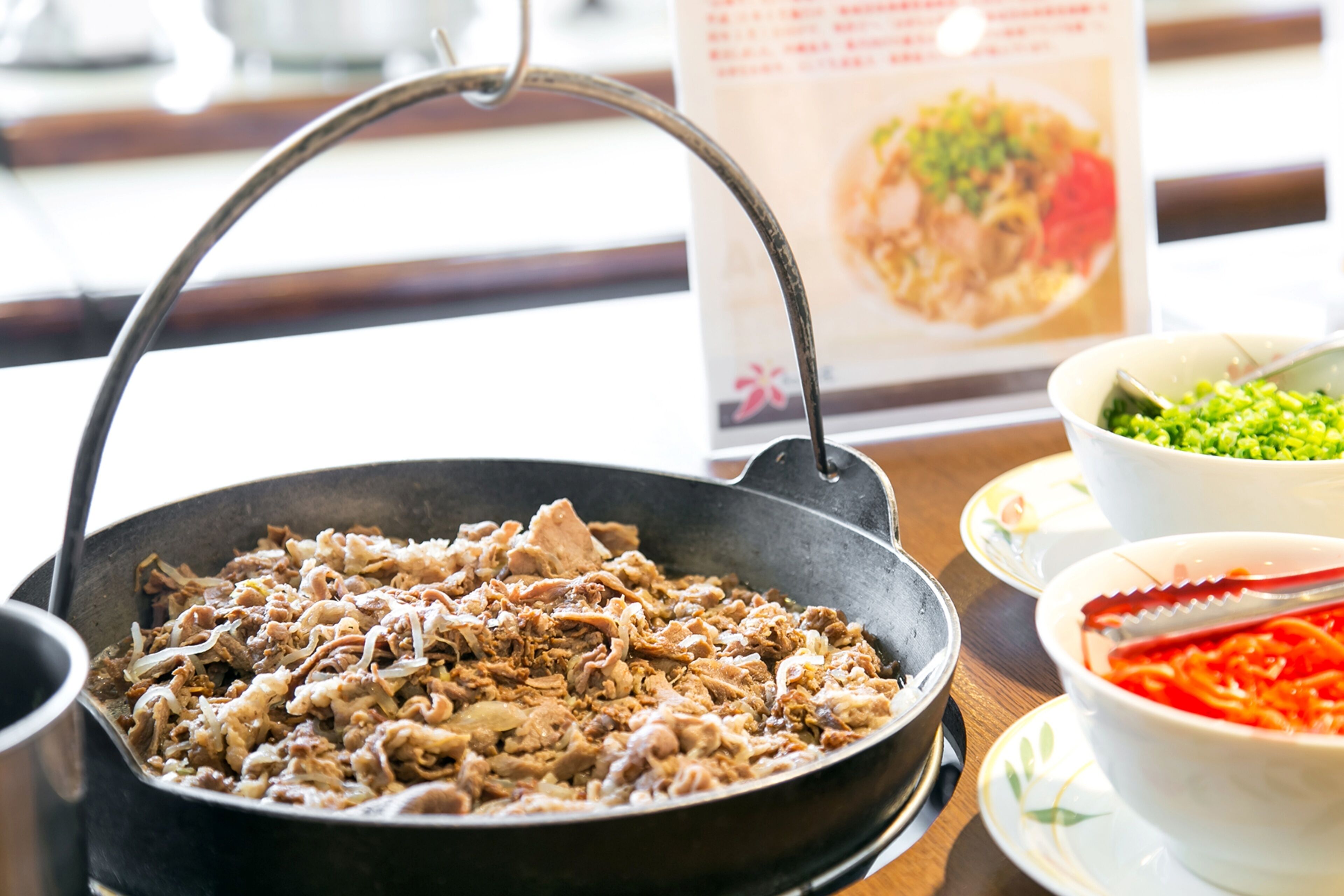 A pan with cooked meat and vegetables, a bowl of green peas, and a bowl of shredded carrots on a table.