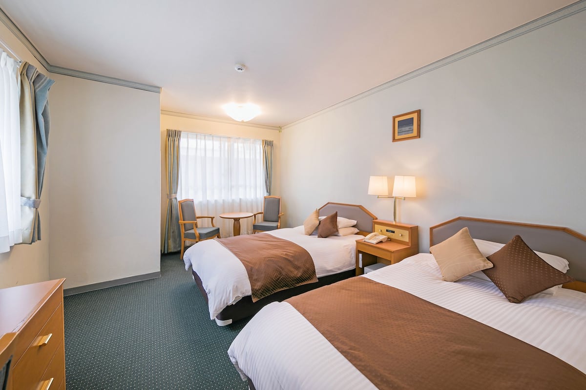 Una habitación de hotel con dos camas, un escritorio, una silla, una mesita y una ventana con cortinas.