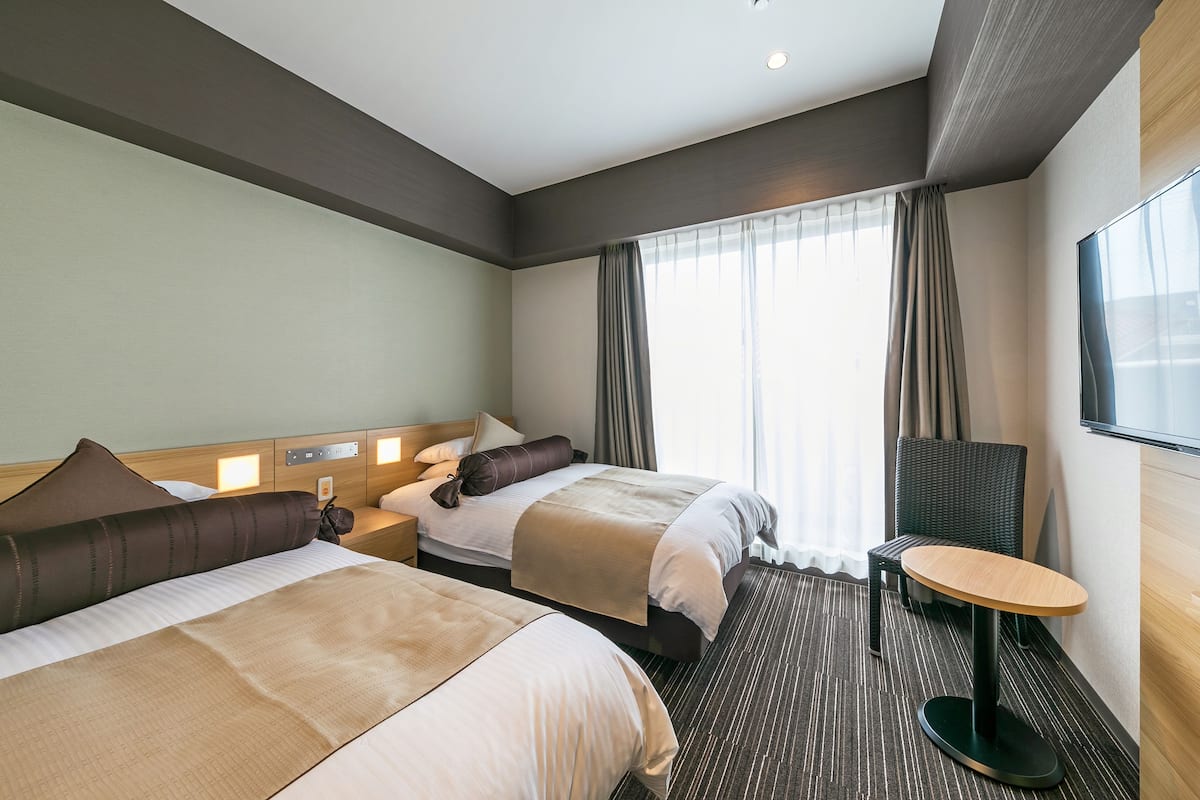 Una habitación de hotel con dos camas, una mesita redonda pequeña y un ventanal grande con cortinas.