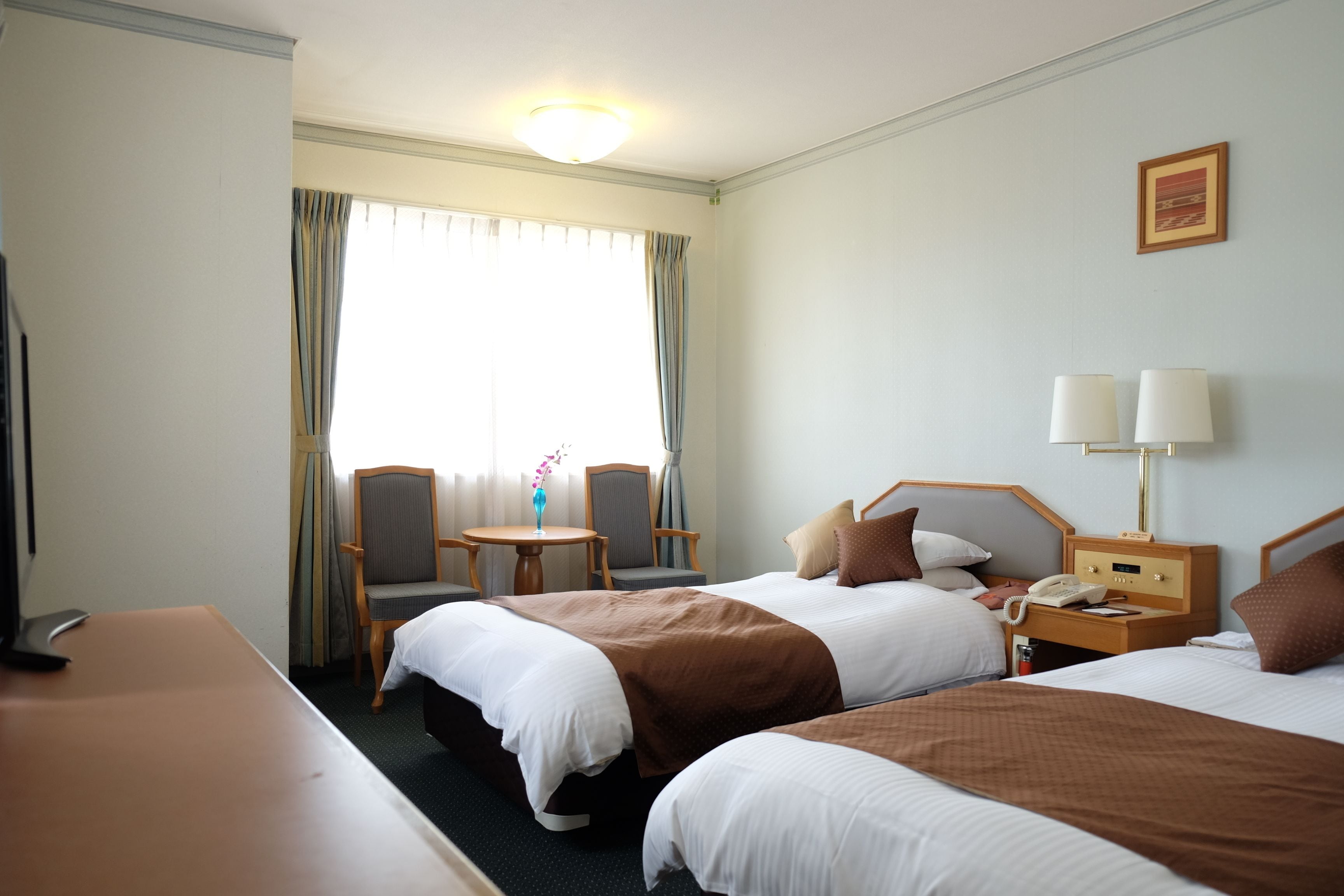Une chambre d’hôtel avec deux lits, un bureau, une chaise, une table, une lampe et une fenêtre avec des rideaux.