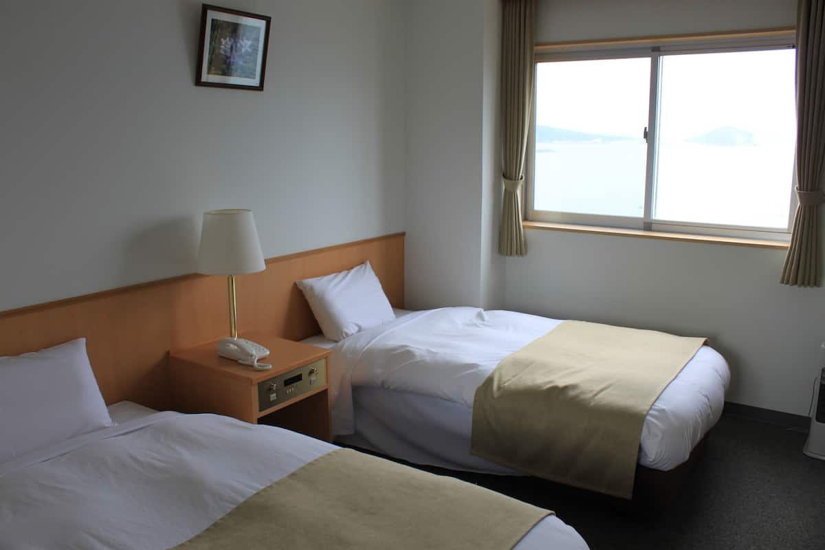 A hotel room with two beds, a nightstand with a lamp, a phone, and a window with curtains.