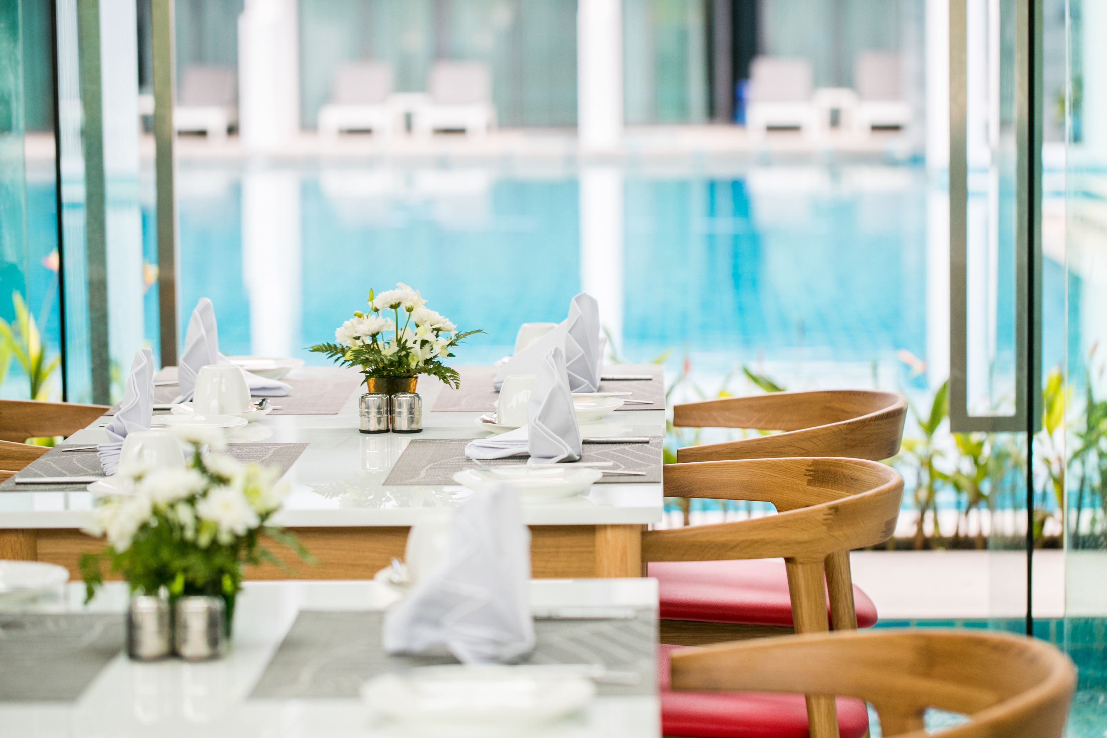 A dining table set for two with white tableware, a vase of flowers, and a view of a swimming pool.