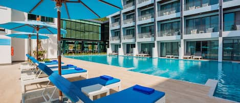 A poolside area with lounge chairs and umbrellas, a modern building with balconies, and a clear blue pool.