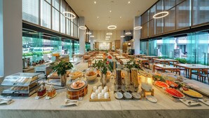 Un comedor amplio con varias mesas y sillas, un bufé con diversas opciones de comida y ventanas grandes que ofrecen vistas al exterior.