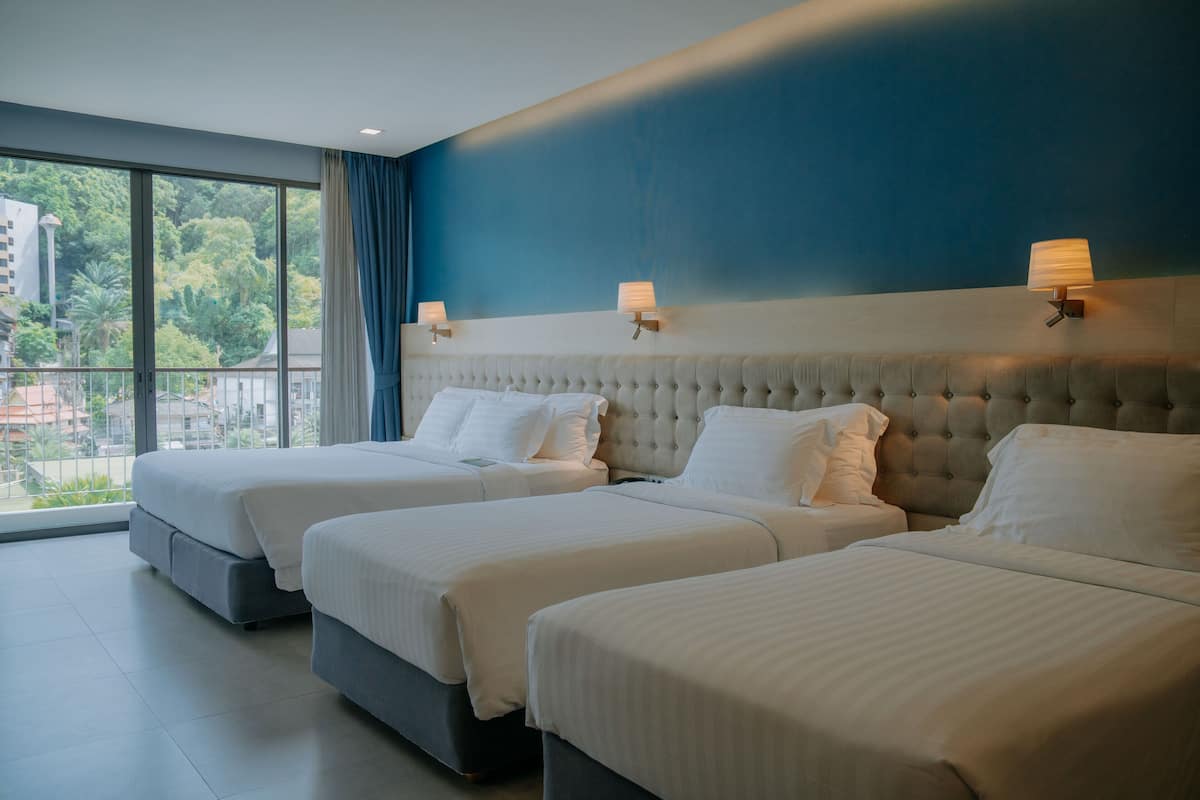 Une chambre d’hôtel avec trois lits, une grande fenêtre et une vue sur la verdure et les bâtiments environnants.
