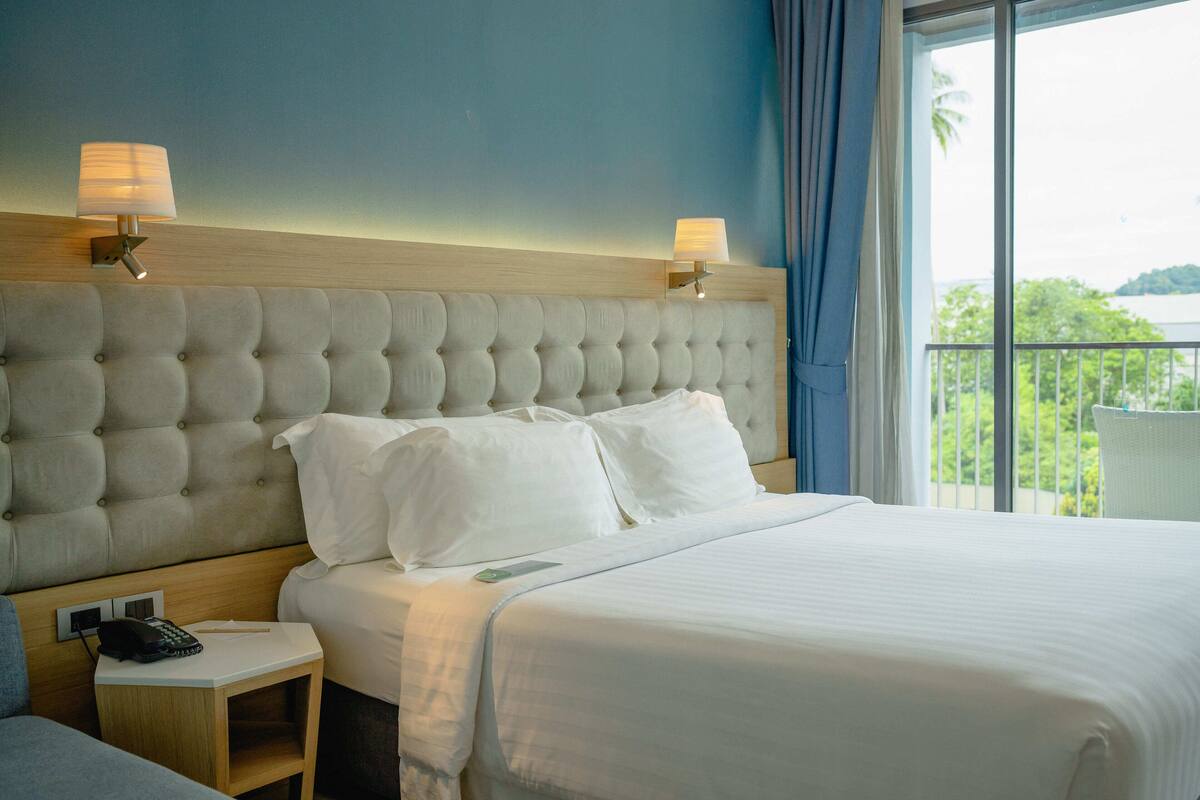 Une chambre d’hôtel avec une tête de lit capitonnée, du linge de lit blanc, une table de chevet avec un téléphone et une vue sur la verdure depuis un balcon.