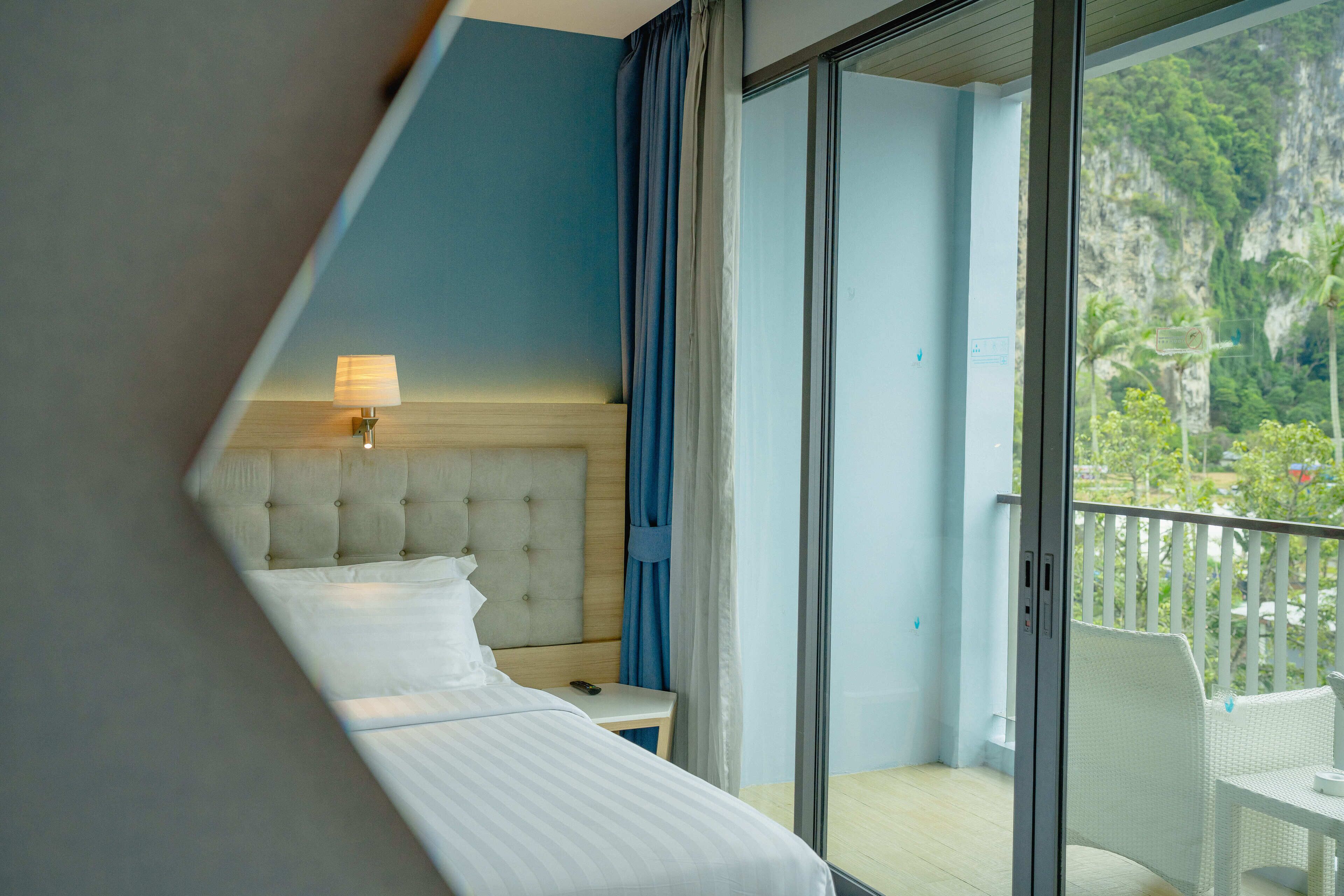 A hotel room with a bed, a nightstand, a lamp, and a view of a balcony with outdoor scenery.