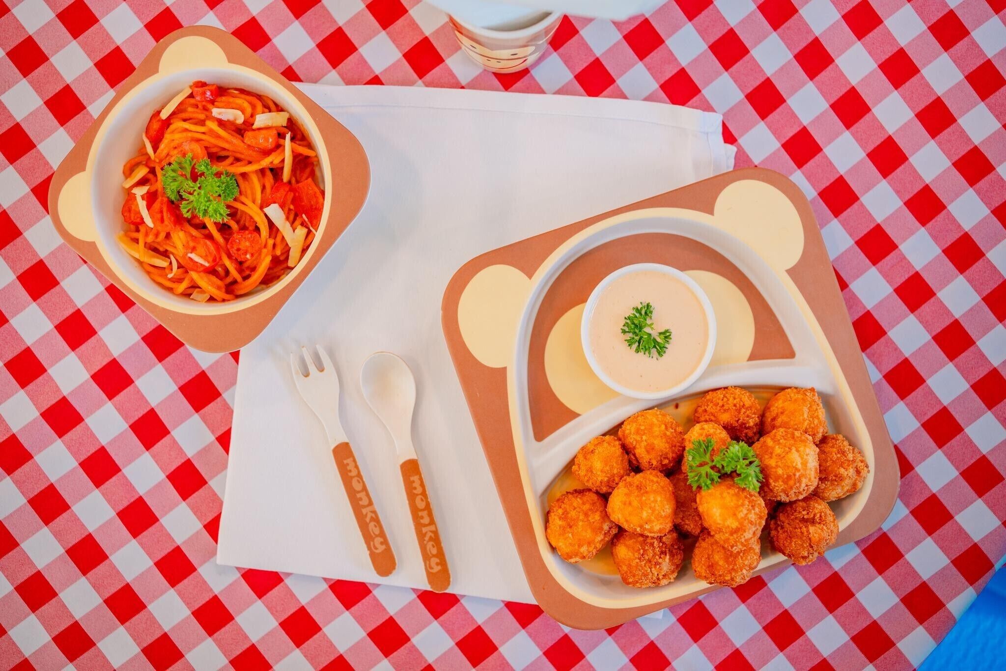 A table with a red and white checkered tablecloth, a bowl of spaghetti with a garnish, a small bowl of sauce, and a container of breaded balls.