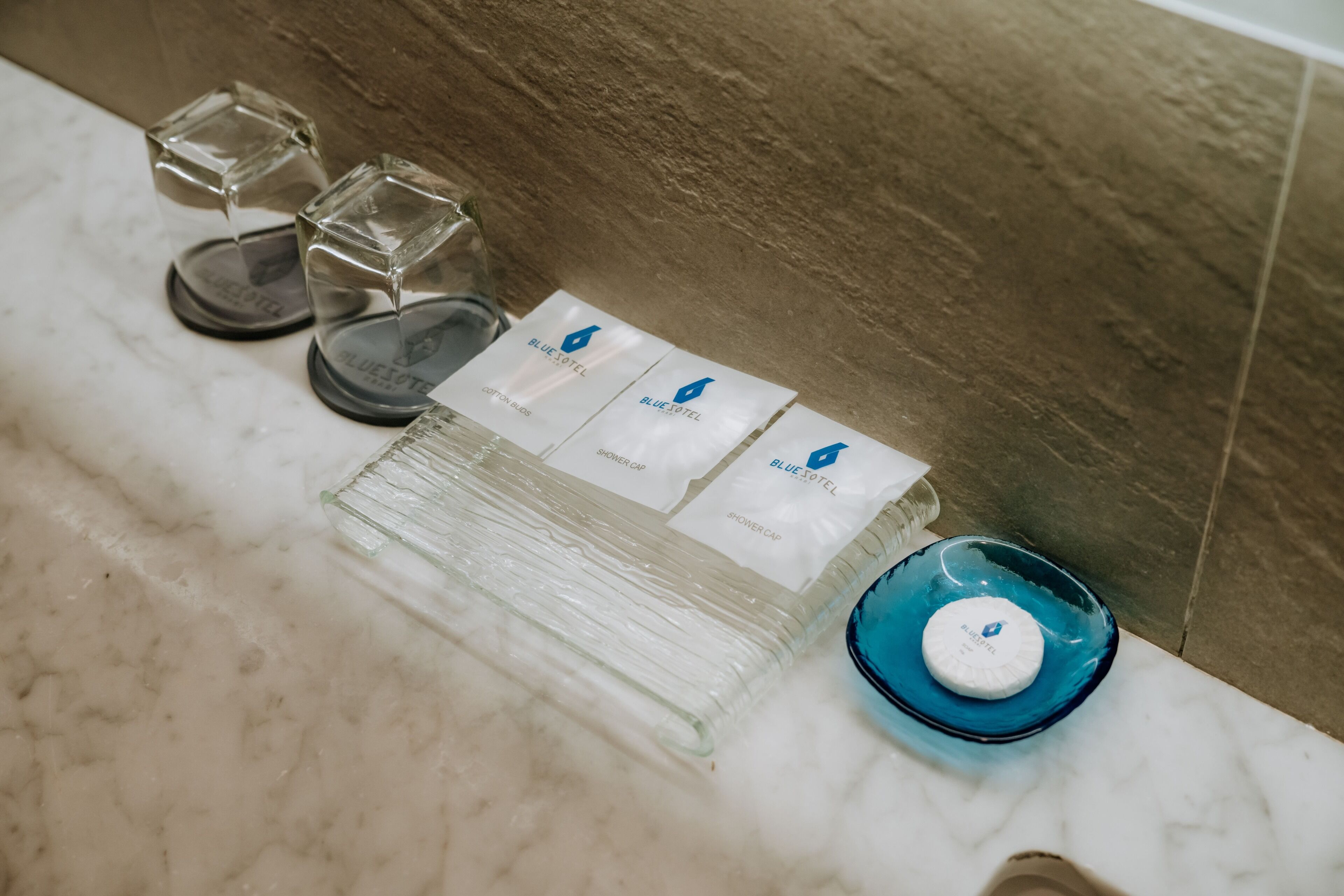 A bathroom with a marble countertop, a blue glass bowl, and several shower caps labeled "Blue Hotel Cotton."