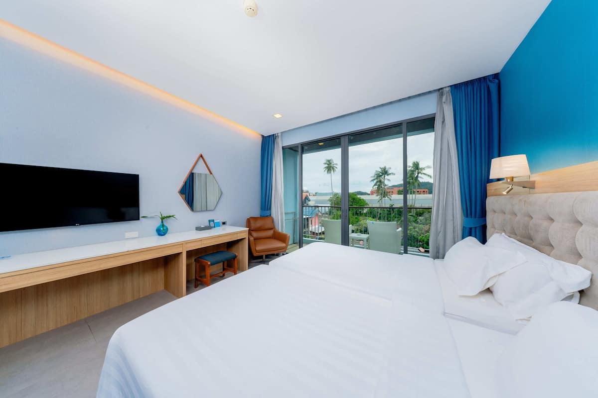 Une chambre d’hôtel moderne dotée d’un grand lit, d’un bureau avec une chaise, d’une télévision à écran plat et d’une grande fenêtre offrant une vue sur les palmiers et les immeubles.