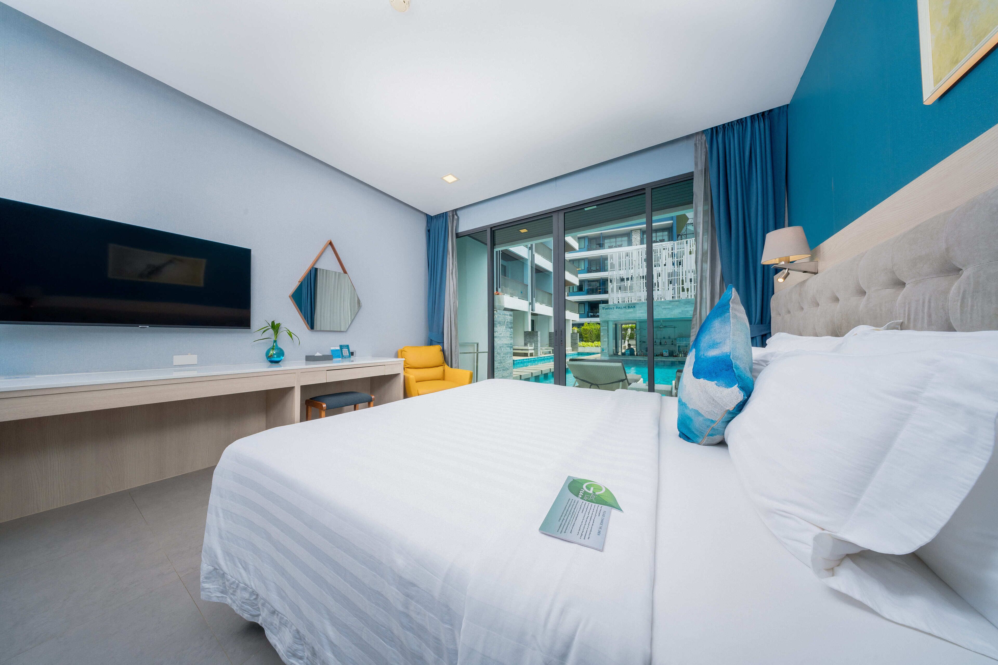 Habitación de hotel moderna con una cama grande, un escritorio, un televisor y vistas a una piscina y un edificio.