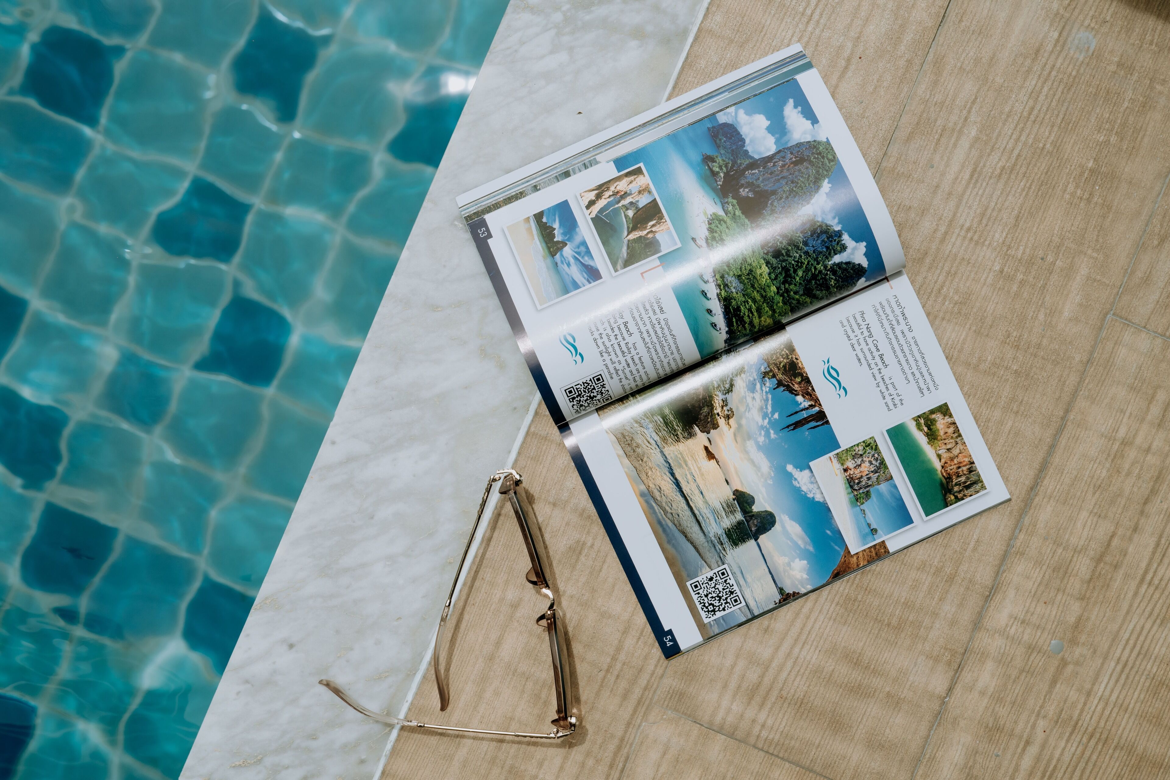 Un magazine contenant des photos de voyage, posé à côté d’une paire de lunettes sur une surface en bois, près d’une piscine.