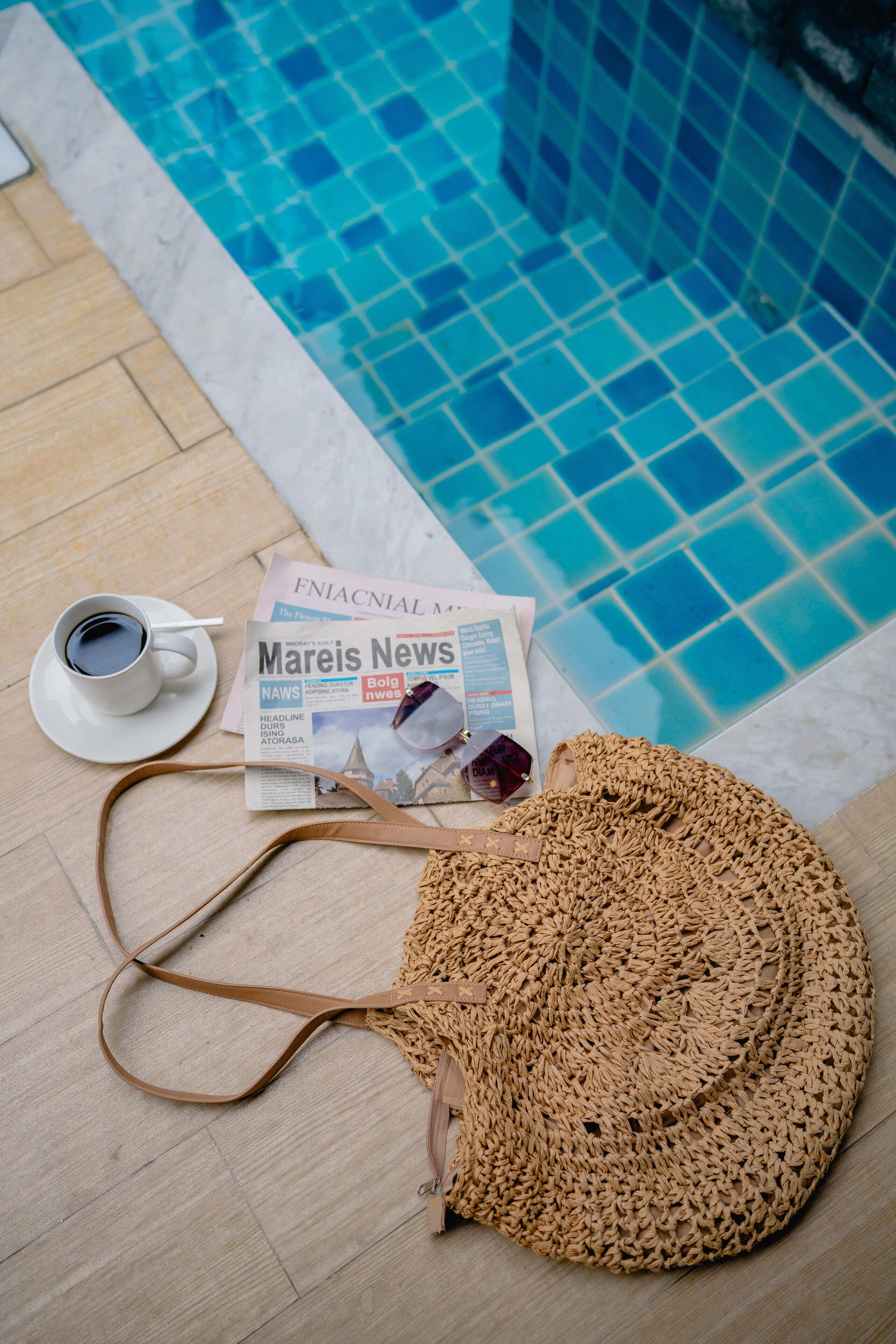 A poolside setting with a newspaper, a cup of coffee, and a woven bag.