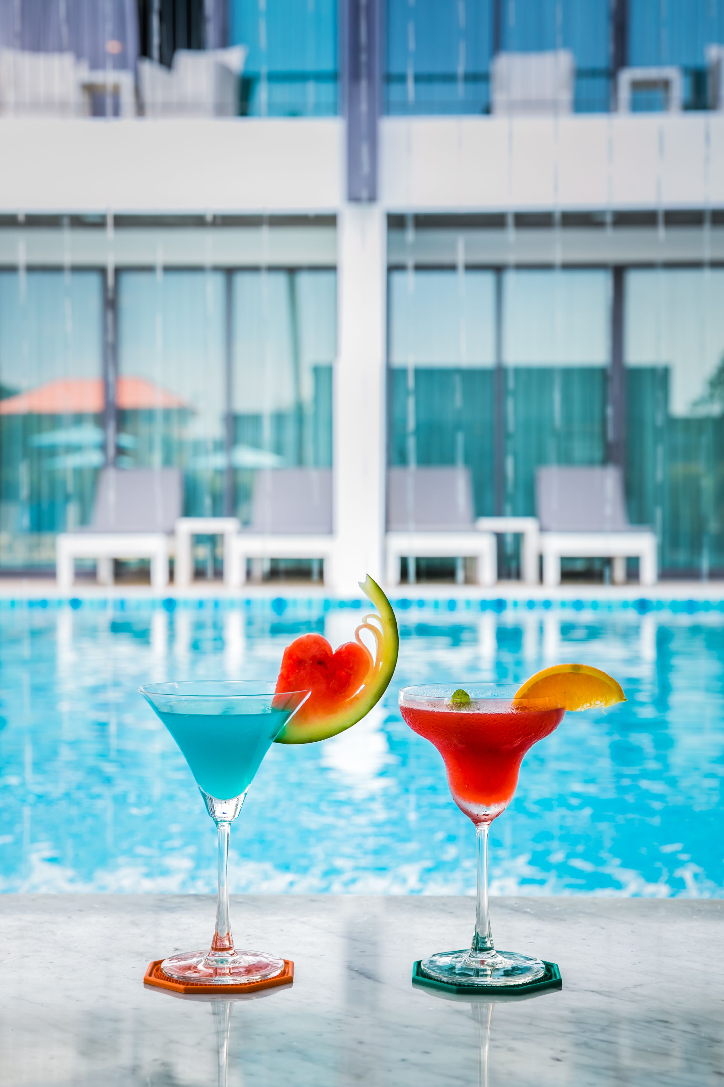 Two cocktails with fruit garnishes in front of a pool with lounge chairs in the background.