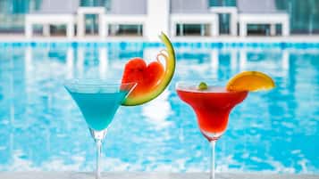 Two cocktails with fruit garnishes in front of a pool with lounge chairs in the background.