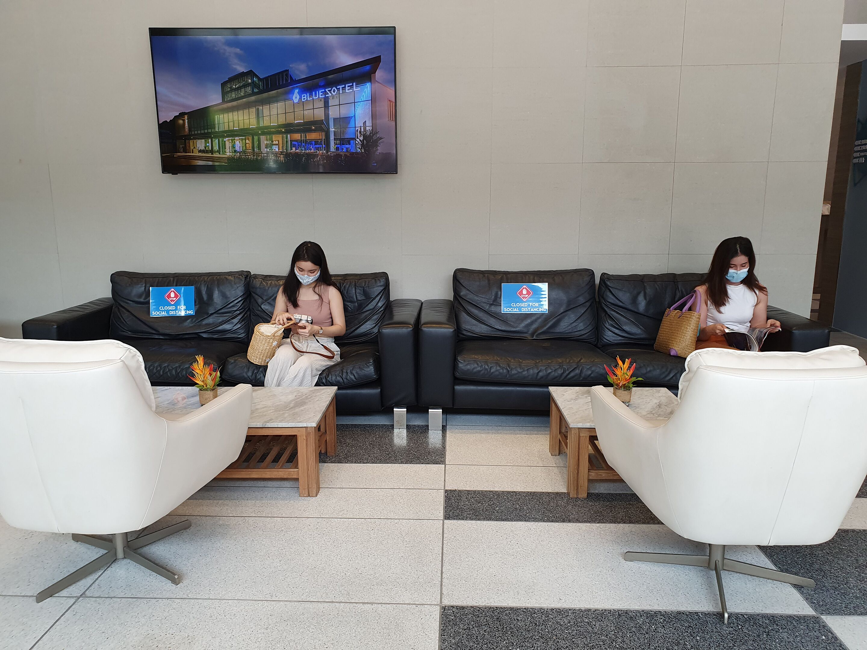 Eine Hotellobby mit zwei schwarzen Ledersofas, einem weißen Sessel und einer Rezeption. Schilder weisen auf Maßnahmen zur sozialen Distanzierung hin.