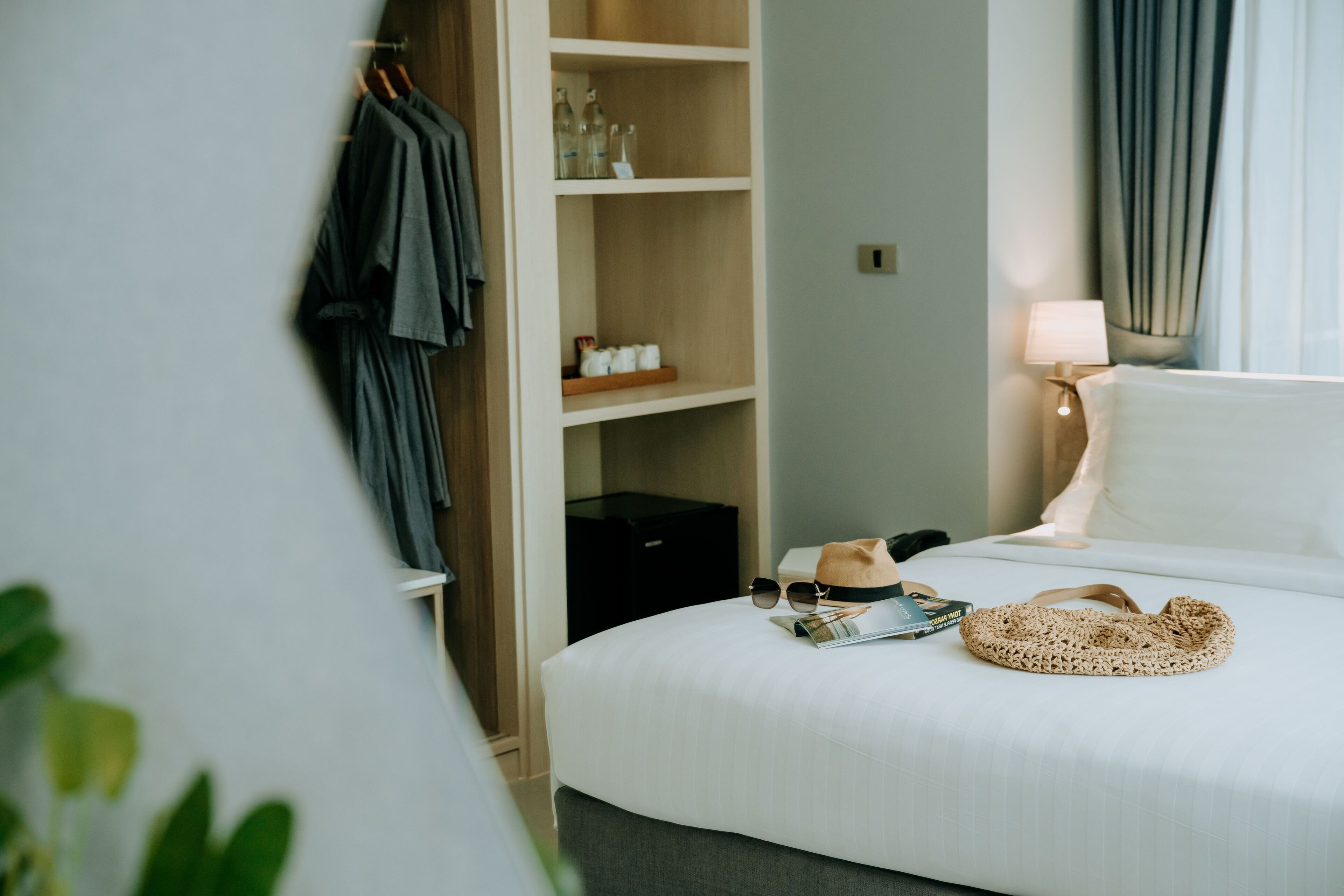 A neatly made bed with a lamp, a hat, and a magazine on it. There is a wardrobe with clothes hanging and a shelf with bottles.