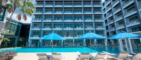 A modern hotel with a large pool area, multiple balconies, and lounge chairs with umbrellas.