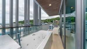 Un balcón con bañera de hidromasaje, barandillas de vidrio y vistas a la ciudad y colinas.
