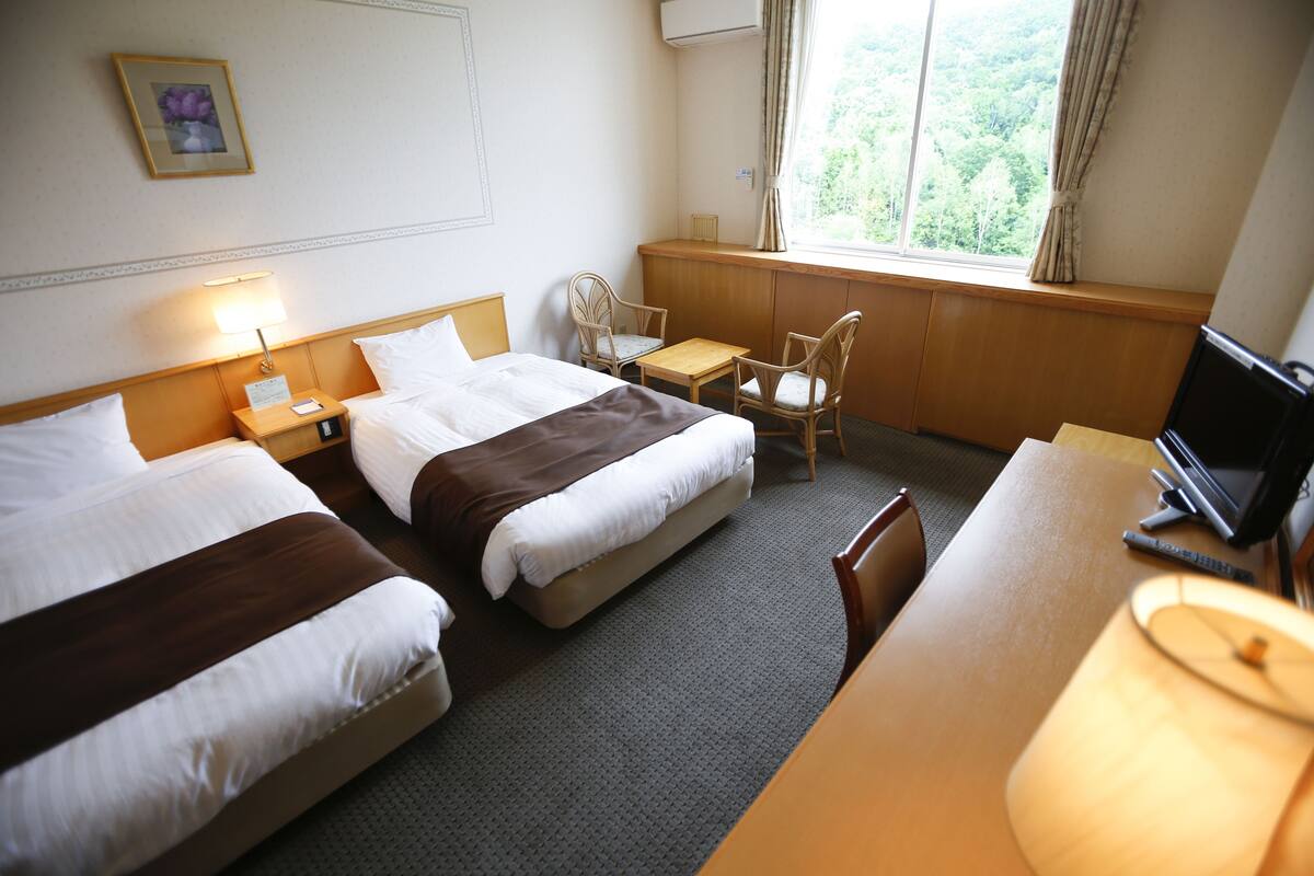 A hotel room with two beds, a desk, a chair, a TV, and a window with a view of the forest.