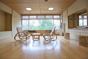 Een ruime kamer met een houten vloer, twee rieten schommelstoelen, een kleine tafel en een groot raam met uitzicht op bomen.