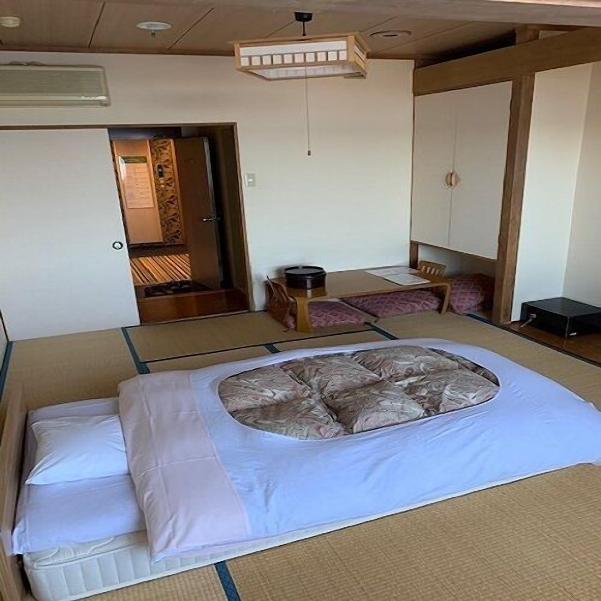 A traditional Japanese-style room with tatami flooring, a low table with a cushion, and a bed with a futon.