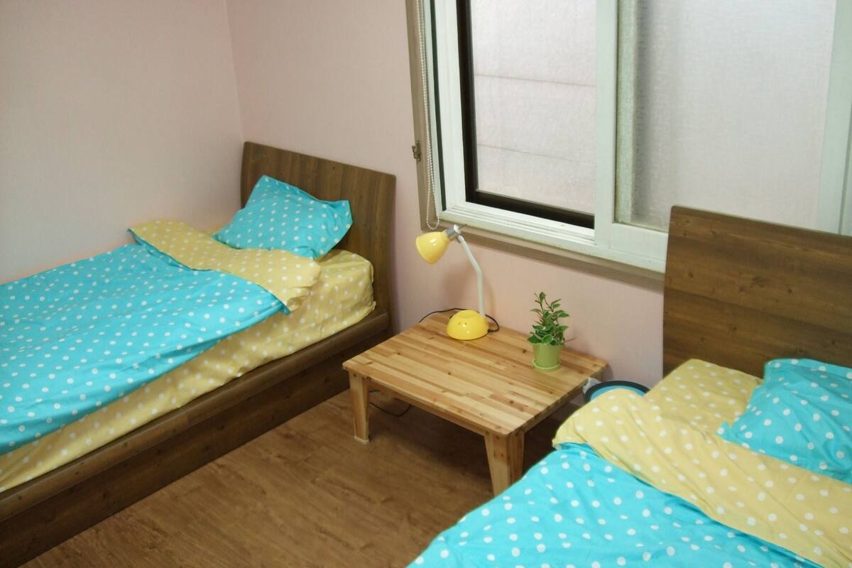 A small room with two single beds, each with blue and yellow polka dot bedding, a wooden nightstand with a lamp, and a small potted plant.