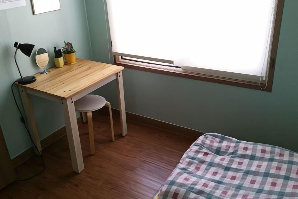 A room with a wooden desk, a chair, a bed with a checkered blanket, a window with blinds, and a small potted plant.