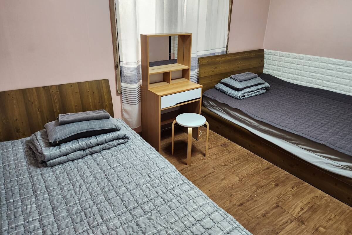 A room with two beds, a wooden desk with a chair, and a window with curtains.