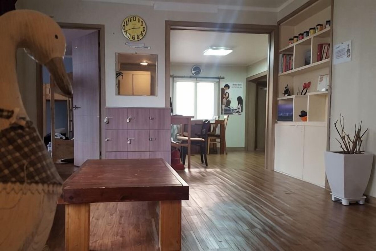 Una habitación con una mesa de madera, un reloj en la pared, una puerta y una estantería con objetos.