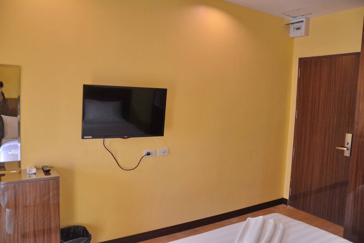 A hotel room with a bed, a television mounted on the wall, a wooden wardrobe, and a trash bin.