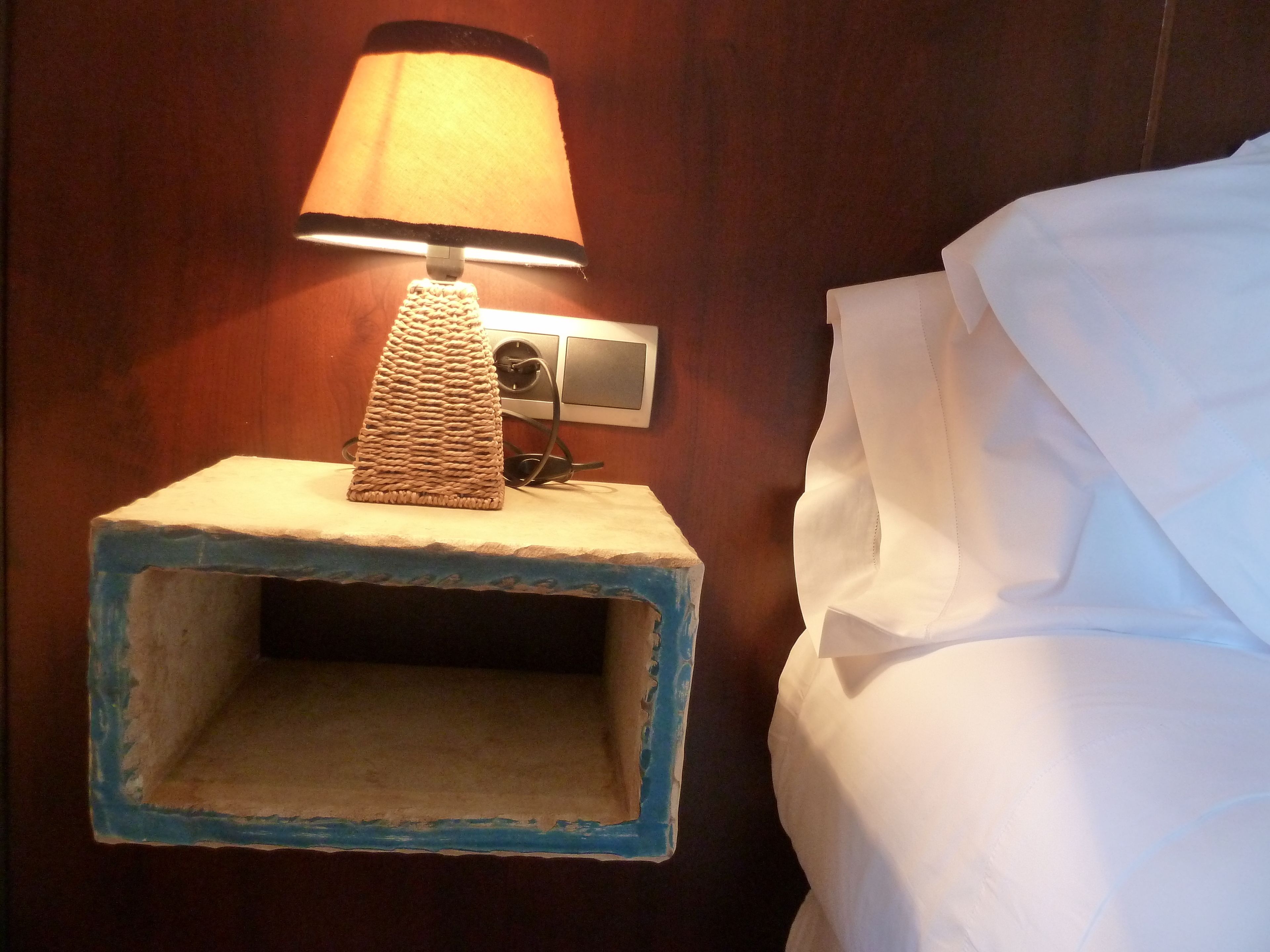 A bedside table with a lamp, a picture frame, and a bed with white sheets.