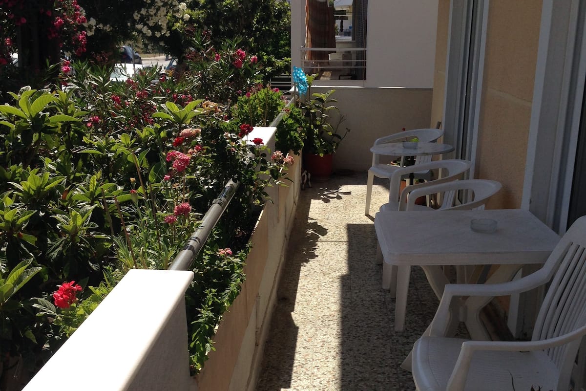 A balcony with white plastic chairs and tables, surrounded by potted plants and flowers.