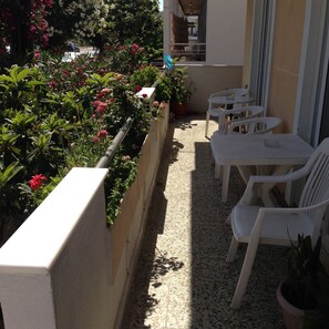 Un balcón con sillas y mesas de plástico blanco, rodeado de plantas y flores en macetas.