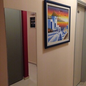 A hallway with a framed painting of a coastal scene, a red pillar, and a door with a number.