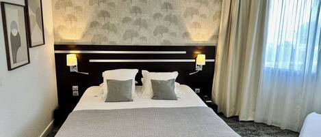 Una habitación de hotel con una cama grande, dos lámparas de noche, un cabecero con estampado de hojas y una ventana con cortinas.