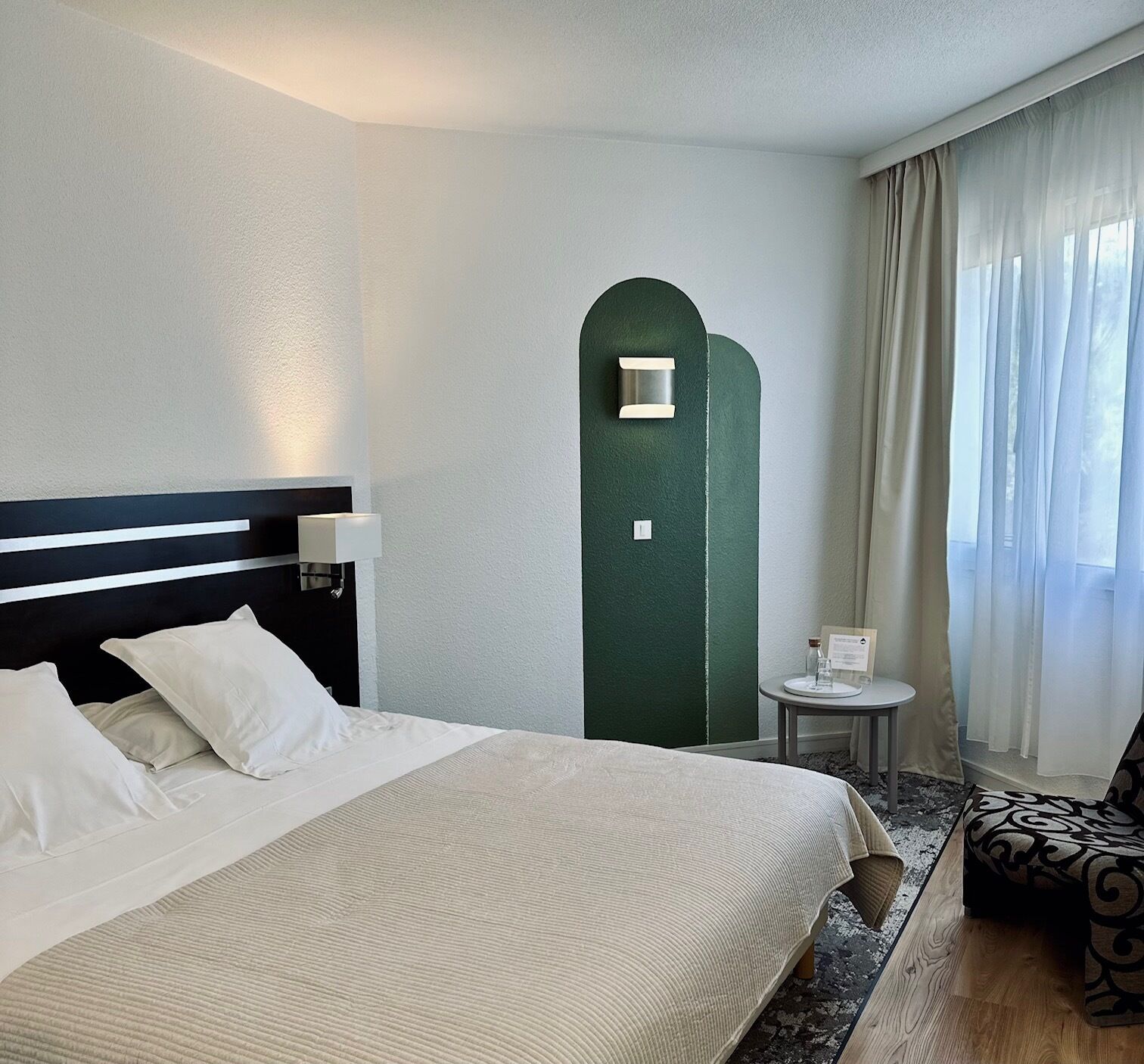 A hotel room with a bed, a bedside table, a chair, a green accent wall, and a window with curtains.