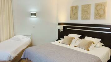 Habitación de hotel con una cama grande, una cama más pequeña, una mesita de noche y una lámpara fijada a la pared. Hay tres obras de arte en la pared sobre la cama.